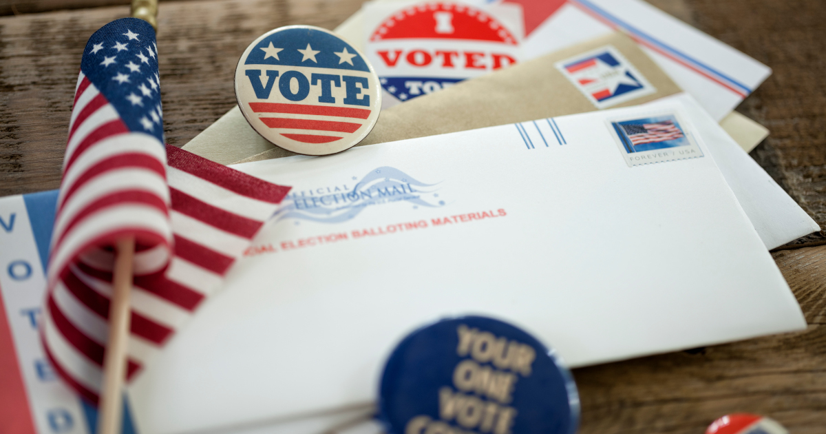 A Small American Flag, Vote Buttons, An Absentee Ballot Envelope, And An I Voted Sticker, All Artfully Arranged Across A Picnic Table Top