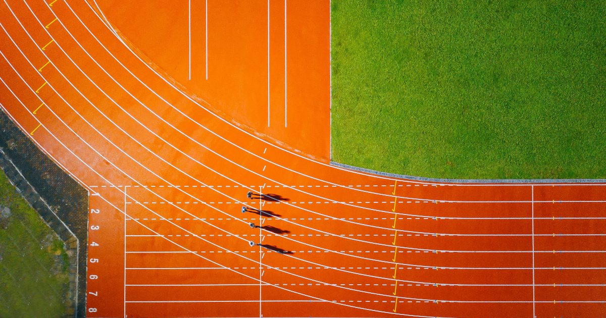 An Athletic Field Seen From Directly Overhead, The Curve of the Orange Cinder Track In The Lower Left Corner And The Green Of The Football Field In The Upper Right Corner, Four Sprinters, Tiny As Ants, Standing Near the Starting Line