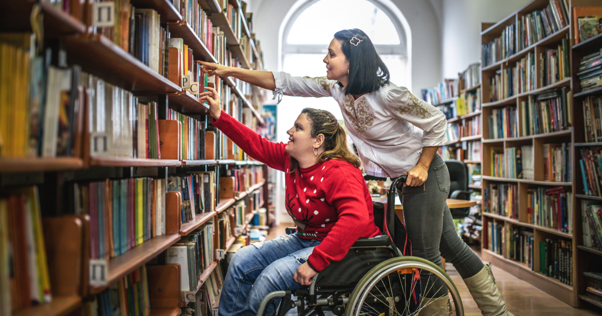 A Young Woman In A Wheelchair Reaching Toward A Book On A Library Shelf While A Young Woman Standing Behind The Wheelchair Reaches To Help Her
