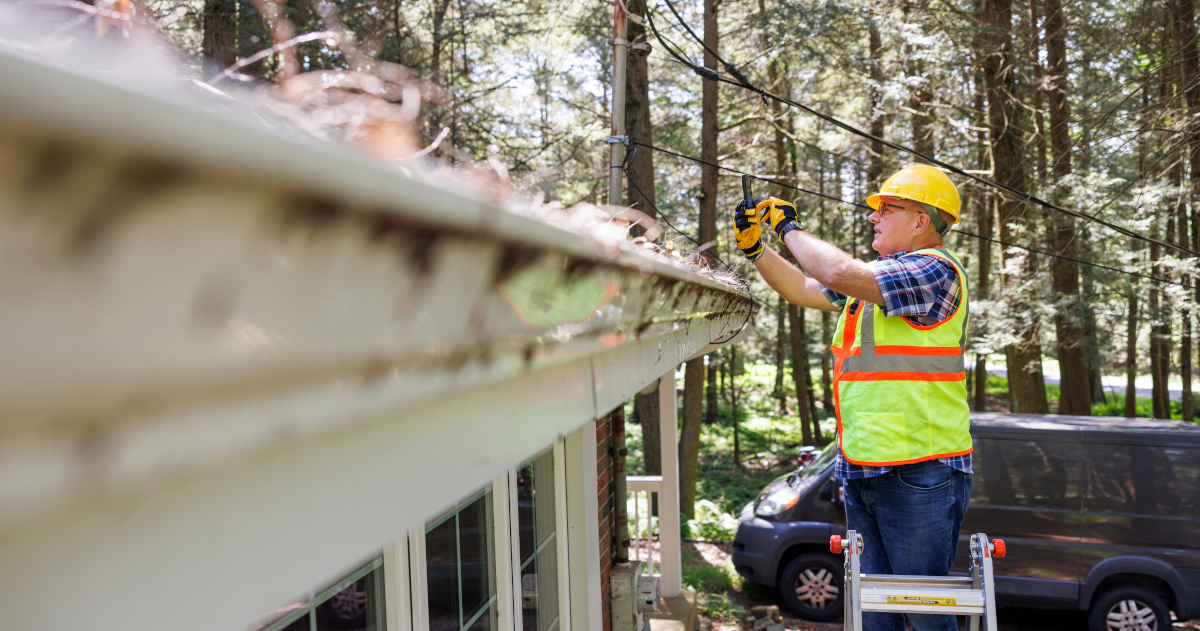 Seen In The Medium Distance, To The Right Of THe Frame, A Middle Aged Man In A High Viz Yellow Vest And Helmet Standing Atop A Ladder, Using His Cell Phone To Snap A Photo Of A Roof, With The Edge Of The Roof Slanting Toward Him