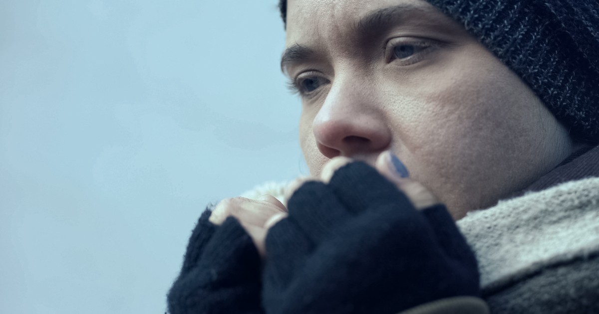 Close Up Of A Woman In A Knit Cap Wearing Fingerless Gloves, Cupping Her Hands To Her Mouth An Blowing On Her Fingers, Against A Wintry Blue-Gray Bakcground