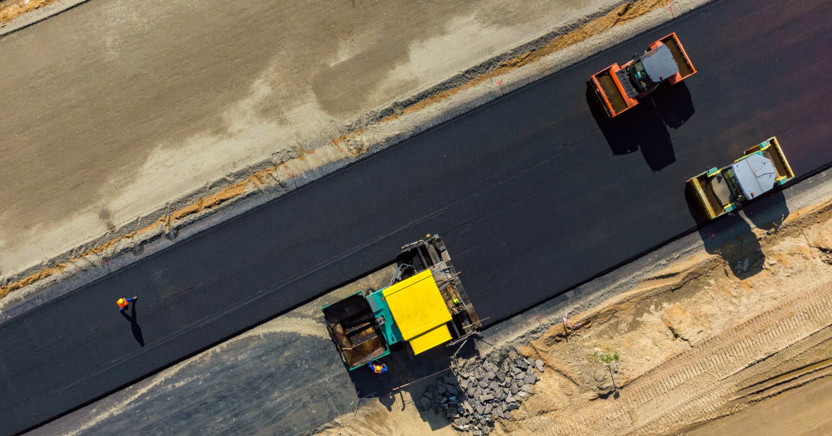 Seen Directly Above From The Air, Three Ashpalht Machines, Two Colored Yellow And One Colored Orange, Smooth A Wide Black Ribbon Of New Road, The Road Surrounded On Either Side by Sandy Dirt Marked By Big Tire Tracks