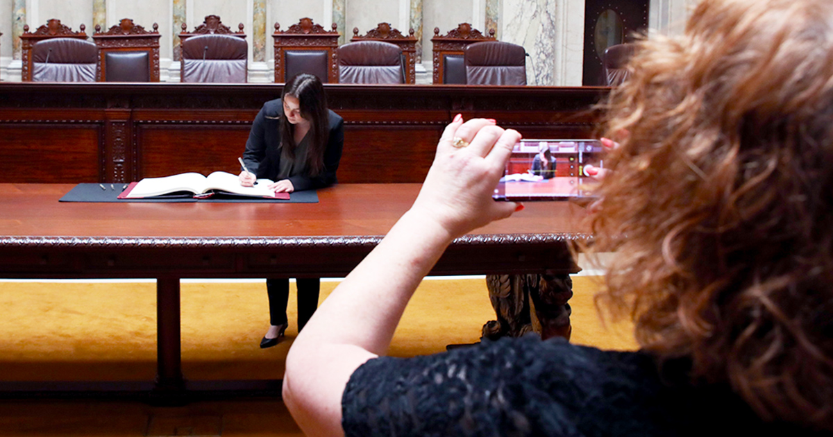 a person bends over a book, writing in it, as another takes a photo