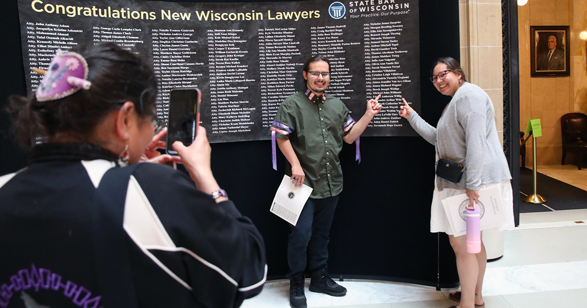 two people point to a name on the name wall as another takes a photo