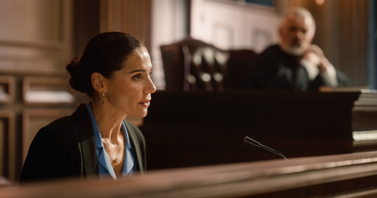 Medium Close Up of A Woman, Dressed In A Dark Suit And With Her Hair Pulled Back, In The Witness Box, With The Judge Out Of Focus To Her Right In The Distance