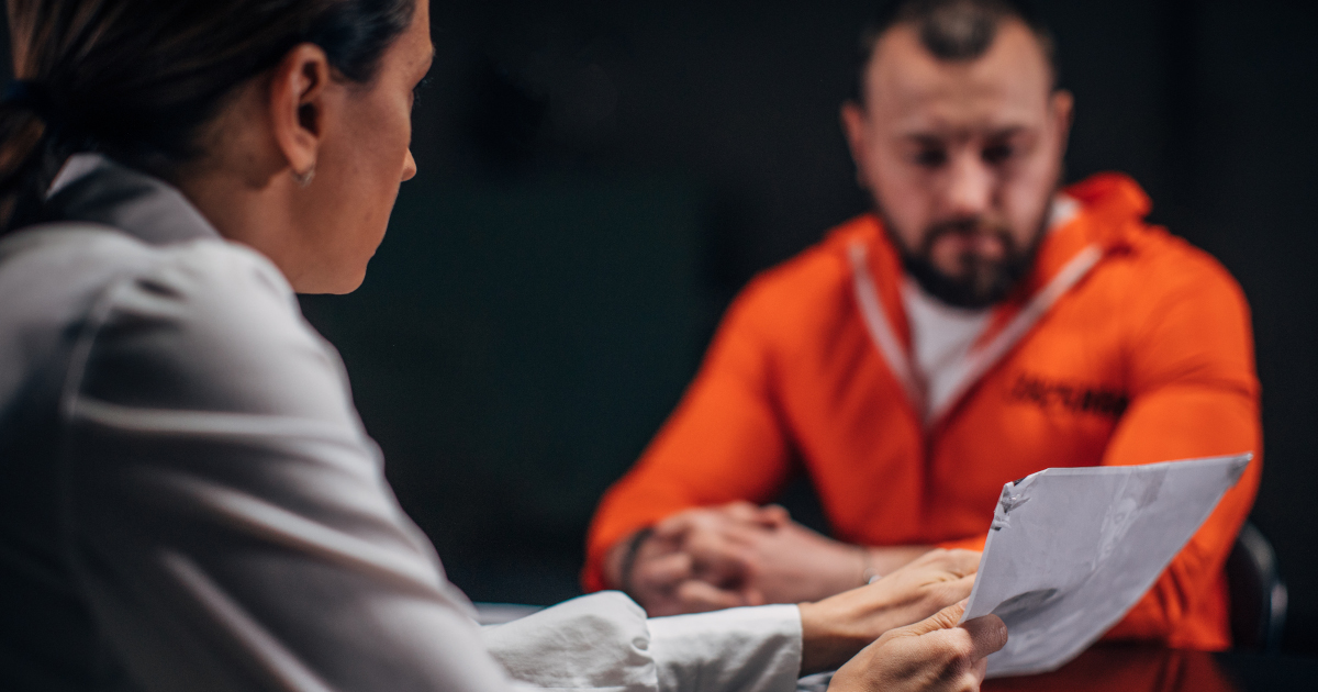 In A Gloomy Room, A Man In An Orange Prison Jumpsuit Sits Across A Table From A Woman In A White Blouse Holding A Piece of Paper Her Right Hand